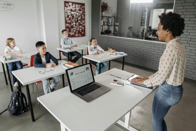 Teacher writing a compelling job post on her laptop while students engage in the background, showcasing a vibrant school culture.