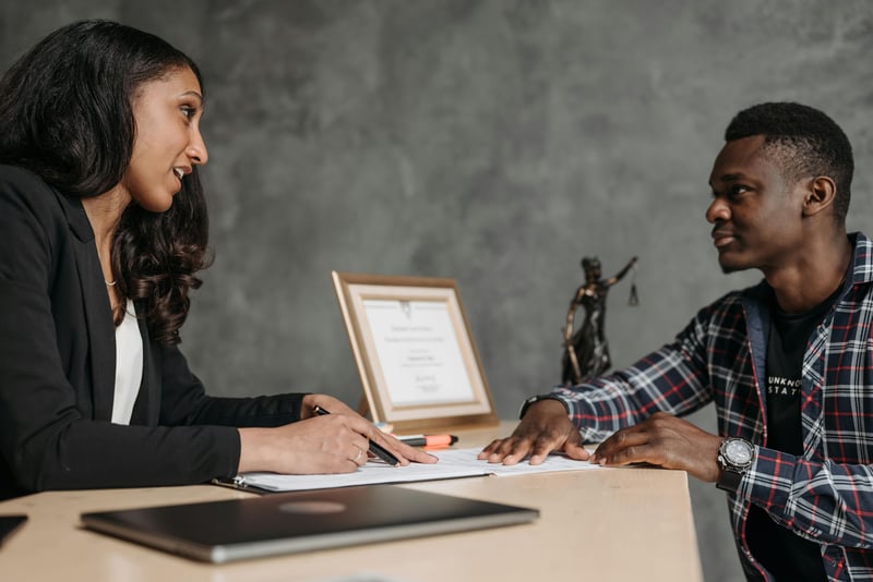 Two teachers sitting across a table from each other and having a discussion. A diploma is framed in the background on the table, and paperwork is laying around the desk.