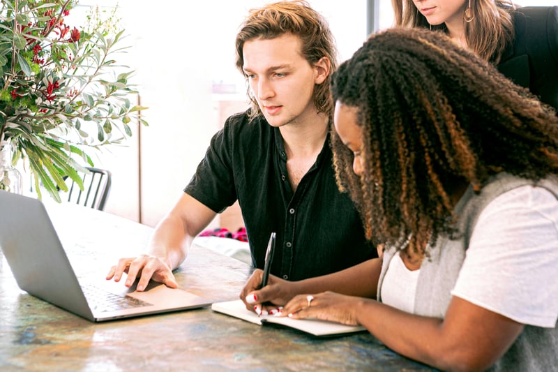 A team of three teachers meeting. One is on a laptop, a second is writing while the third is standing behind them looking at the screen.