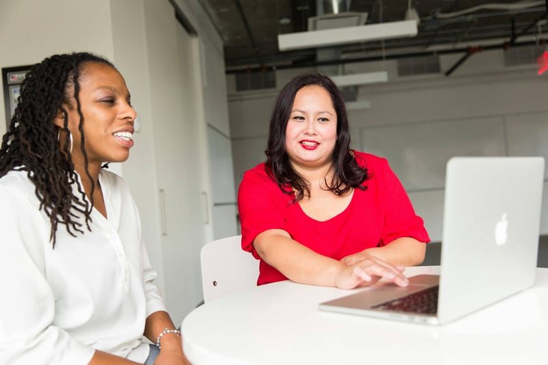 Two teachers in an office are working on a laptop. One of them is smiling.