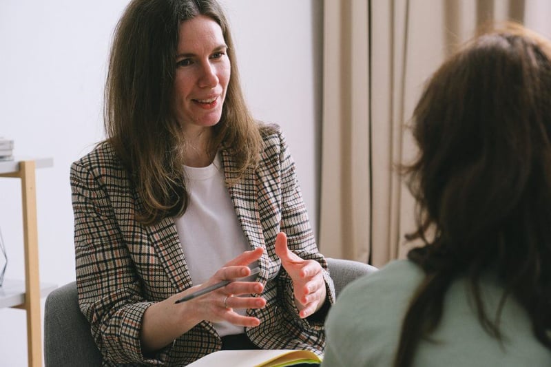 Two teachers facing each other with one talking to the other, as if she was explaining a specific topic. She is holding a pen in her right hand, and has a notebook on her lap.