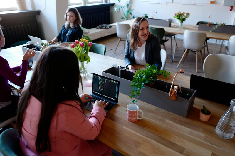 A team of 5 teachers are sitting on a desk in what looks to be a coworking session. Some have laptops in front of them, and coffee mugs. Two are in discussion, while a third is laughing.