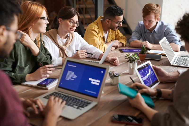 A team of seven teachers are meeting around a table discussing and planning what seems to be an onboarding program. Some laptops are open on slide presentations.