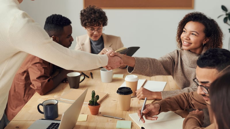 Six teachers are around a conference table. Some are working together, while two are shaking hands across the table from each other showing the positive impact of an effective onboarding.