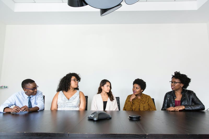 Five teachers are all sitting on the same side of a conference table, facing the camera. they are looking at each other. The seem to be sitting in a conference room.