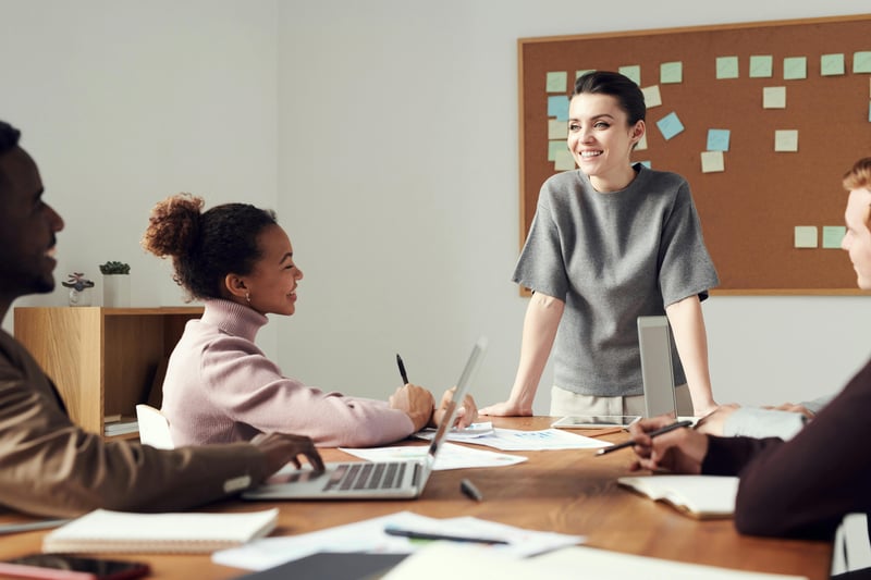 A team of multiple teachers are sitting at a conference table facing one standing person who seems to be welcoming them for a sort of orientation and onboarding. The table is littered with notebooks, papers, and pens.