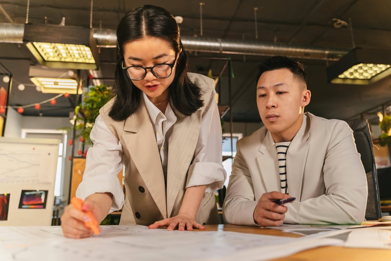 Two teachers are side by side on a desk. One of them is standing up as she's highlighting lines on a big paper note while the other is observing her work.