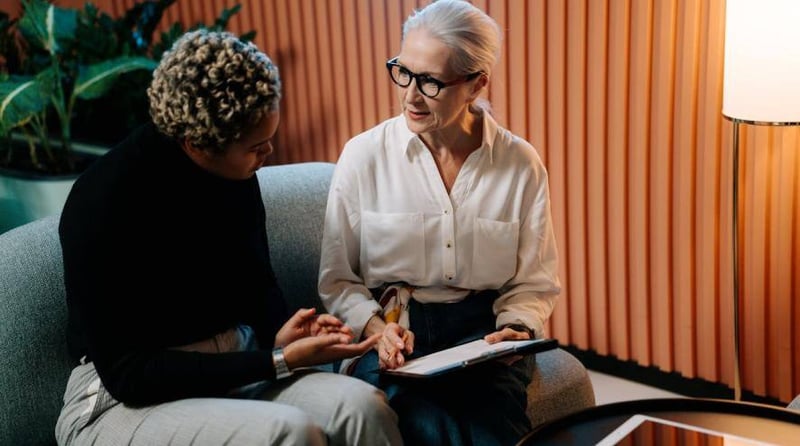 Two persons sitting on a couch are in a discussion. One of them is holding a clipboard.