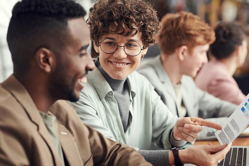 Two teachers are sitting side by side and having a discussion while one of them is smiling. In the background we see other teachers having meetings.