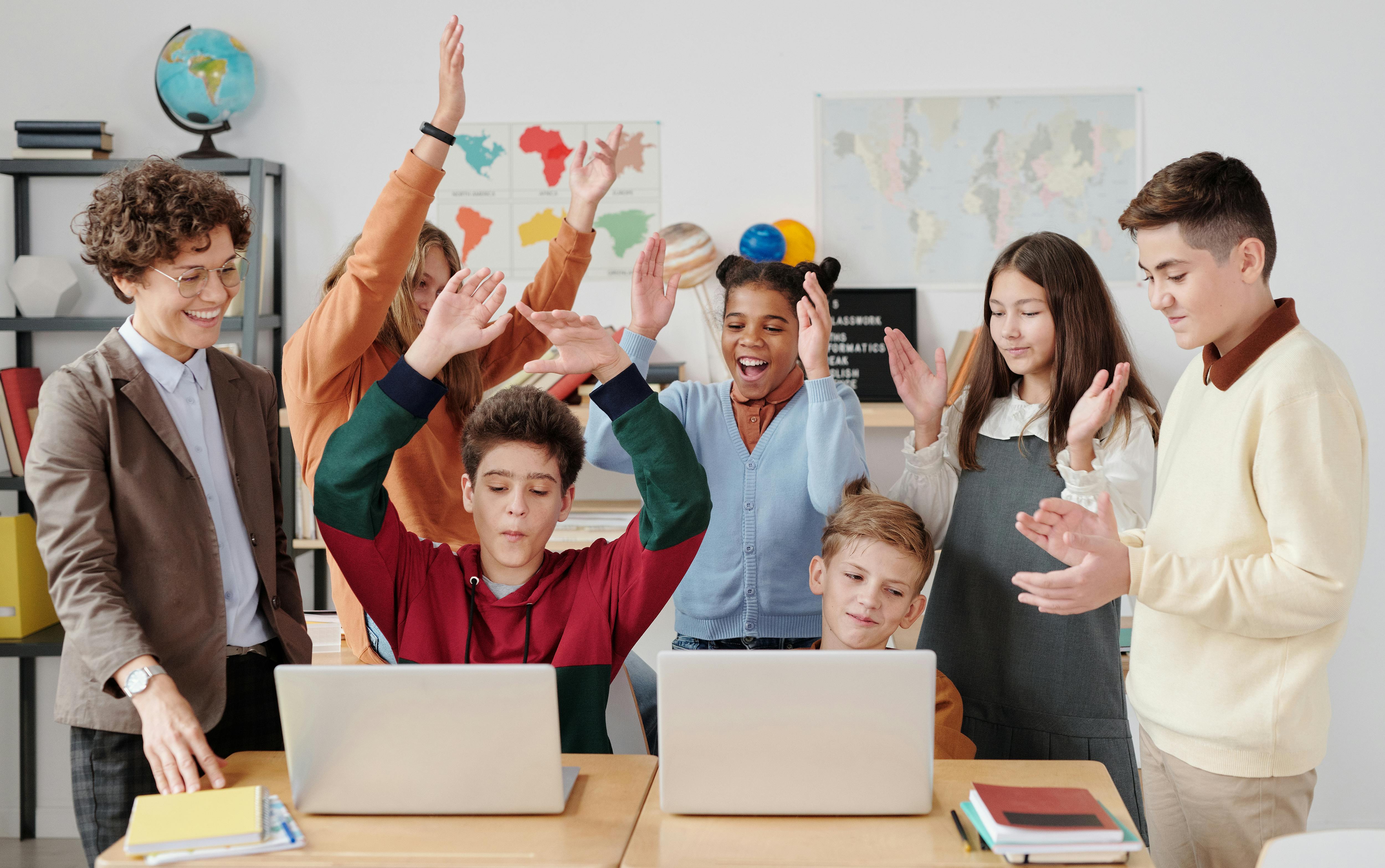 A class of young students gathered around two laptops celebrating a success while a teacher looks over them