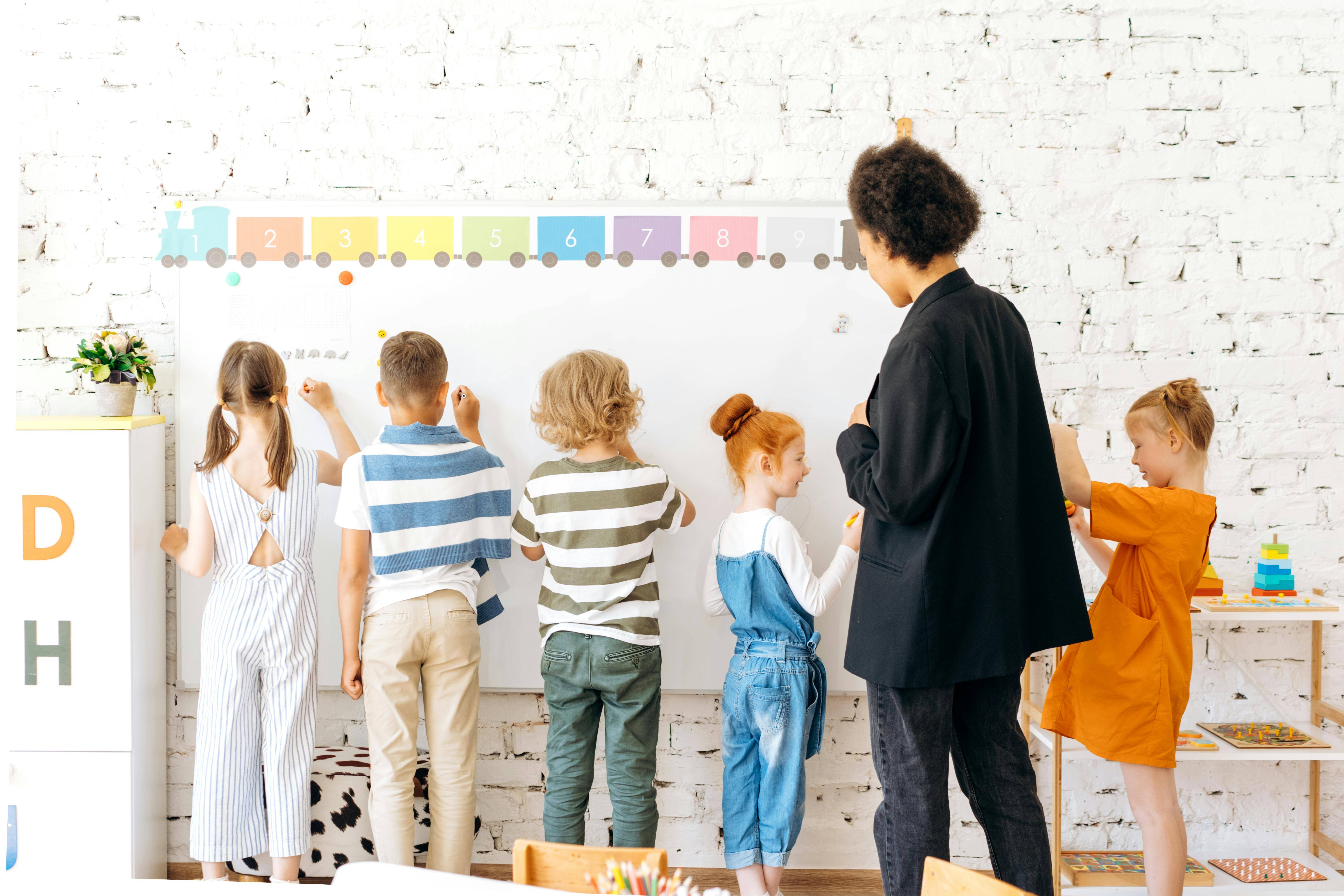 Five young students writing on a whiteboard with a teacher supervising them