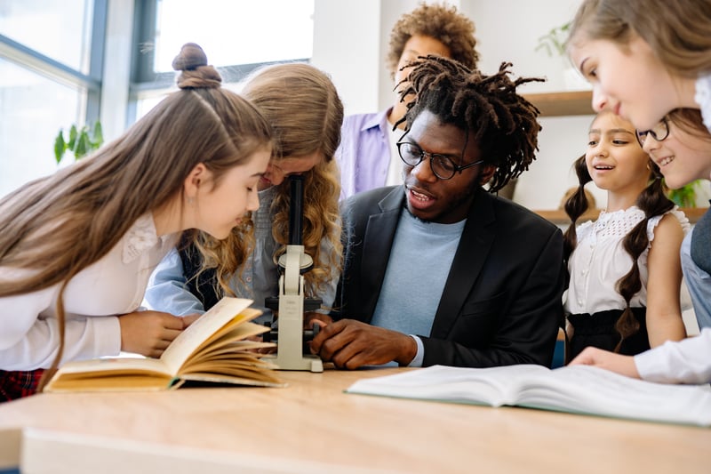 male teacher reading with smiling elementary students in a classroom, representing inclusive teaching environments and the importance of educator support to prevent teacher burnout.